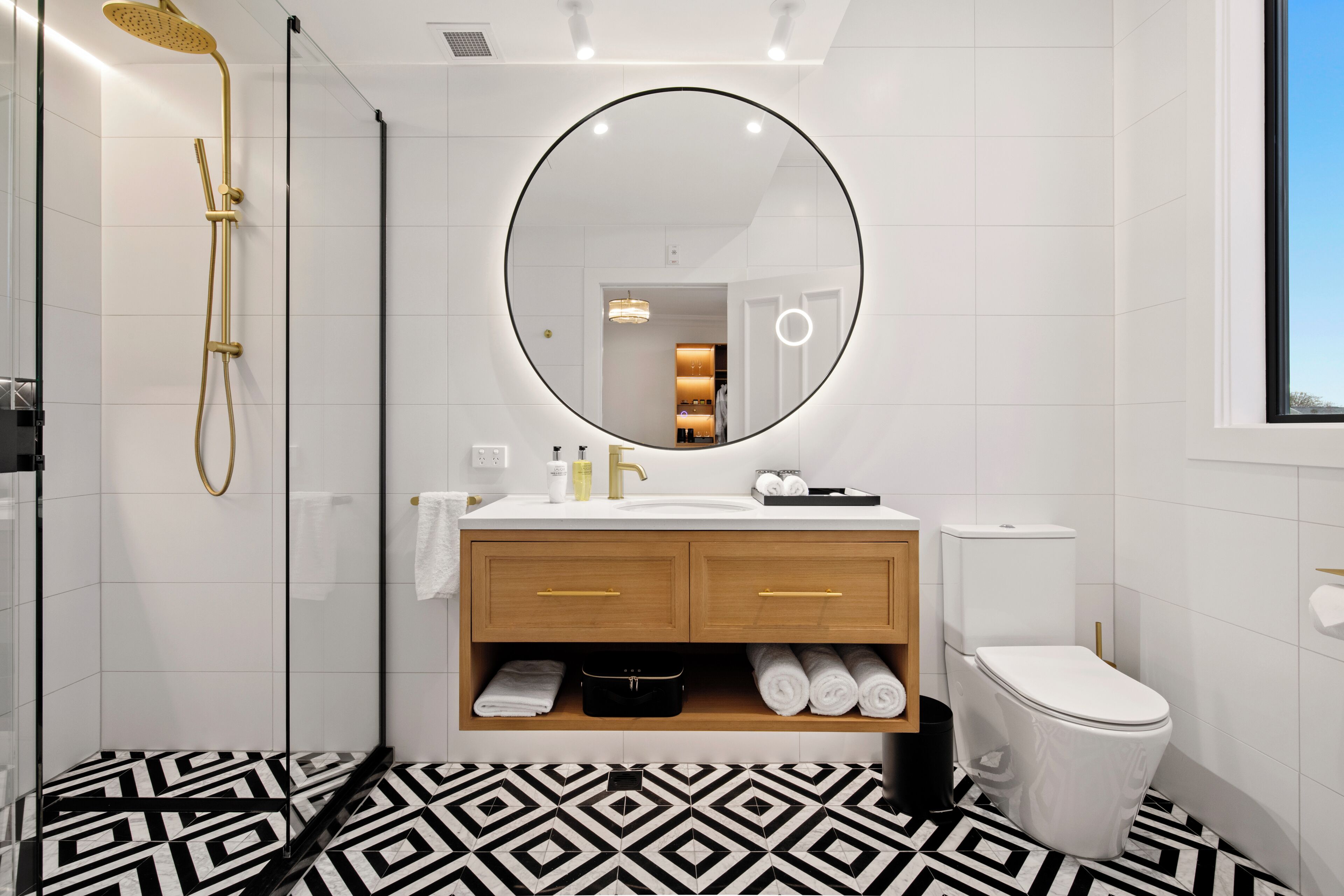 A modern bathroom with a glass-enclosed shower, a round mirror, a white sink with wooden cabinets, and a toilet.