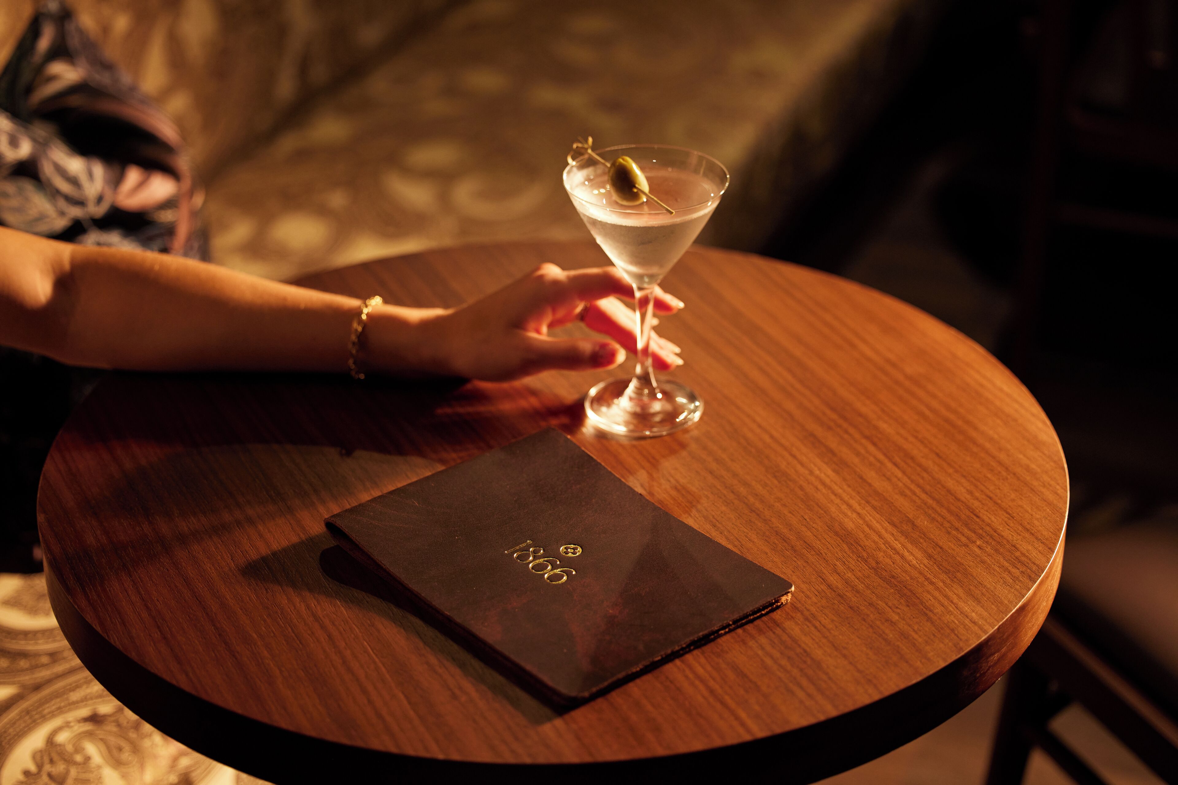 A hand holding a martini glass with a lemon twist on a wooden table. A black booklet with gold lettering is placed on the table.