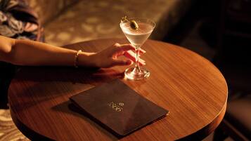 Una mano sosteniendo una copa de martini con una rodaja de limón sobre una mesa de madera. Un librito negro con letras doradas está sobre la mesa.