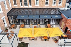 An outdoor seating area with yellow umbrellas labeled "Clements Hotel," featuring wicker furniture and a brick building in the background.