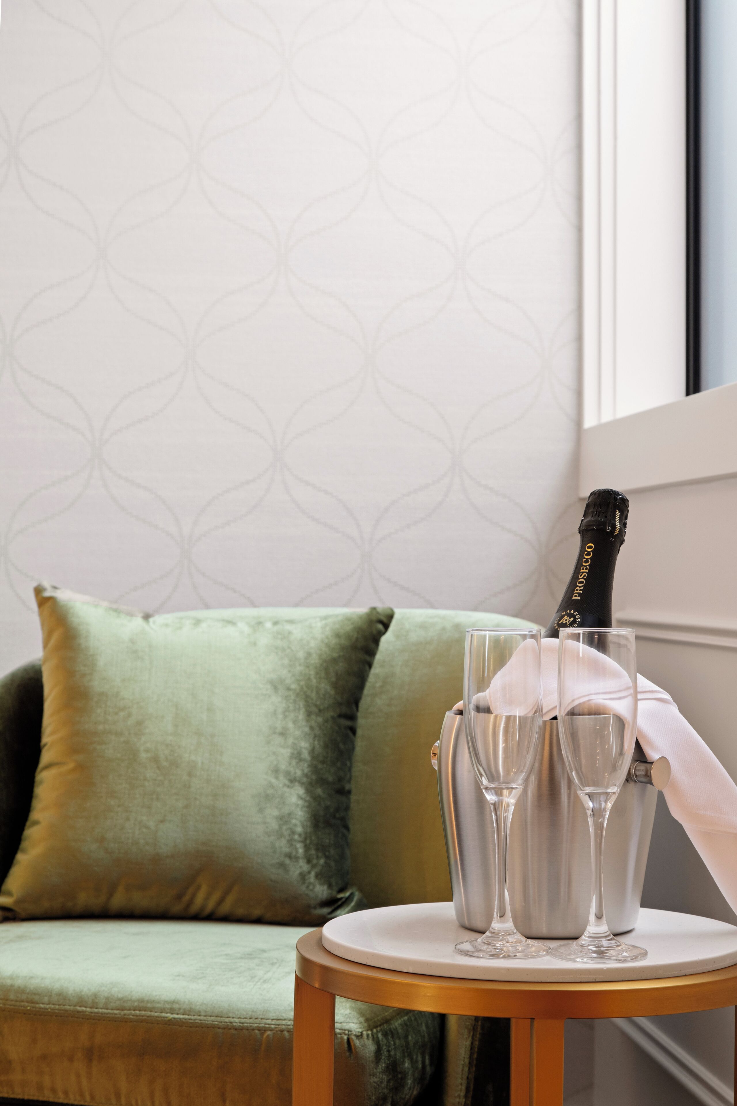 A room with a green sofa, a small round table with two champagne flutes and a bottle, and a patterned wallpaper.