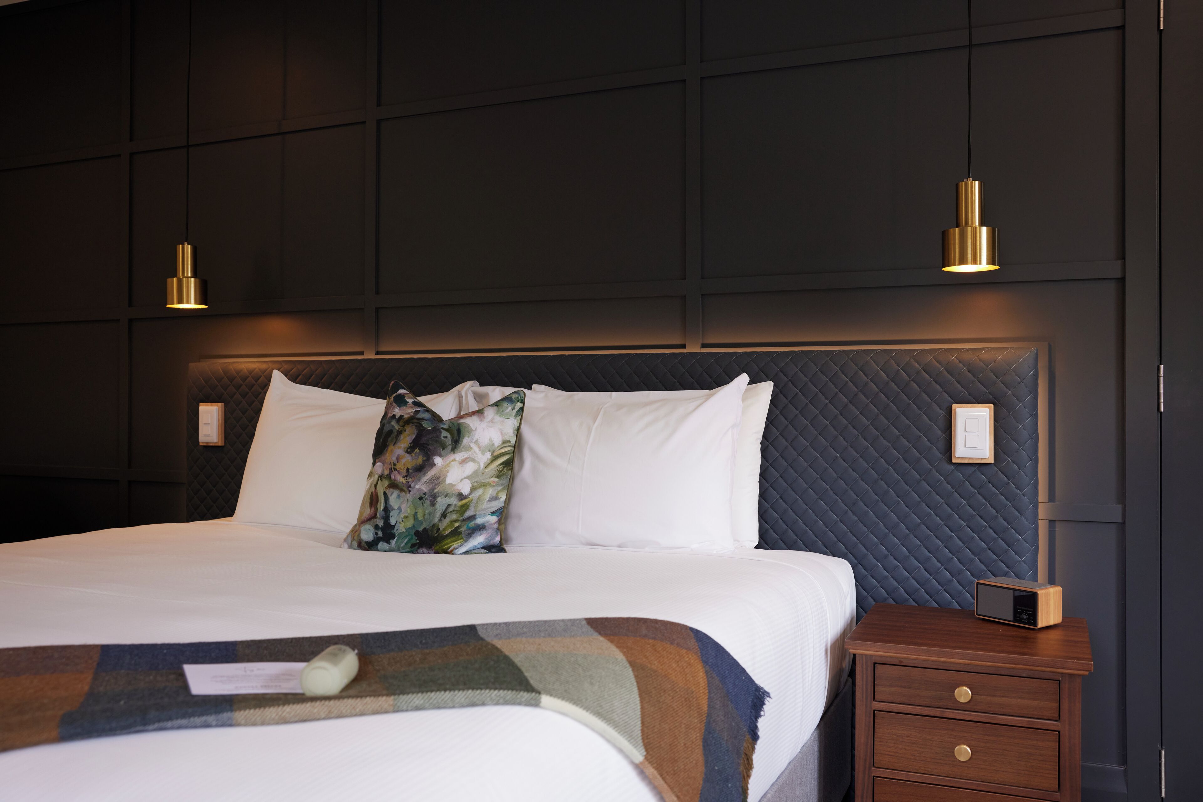 A neatly made bed with a quilted headboard, two bedside lamps, a wooden nightstand with a radio, and a wall-mounted light switch.