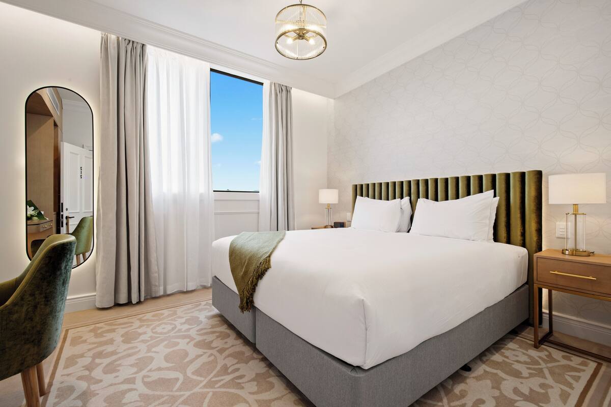 Ein Hotelzimmer mit einem großen Bett, einem Nachttisch mit Lampe, einem Sessel und Blick ins Freie durch ein Fenster.