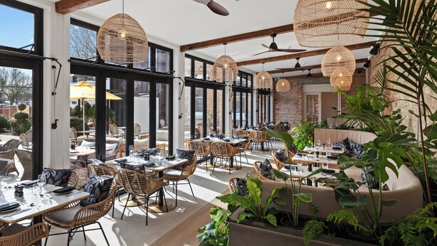 Un espace de restauration intérieur aménagé avec des meubles en osier, de grandes fenêtres et des lampes suspendues en osier.