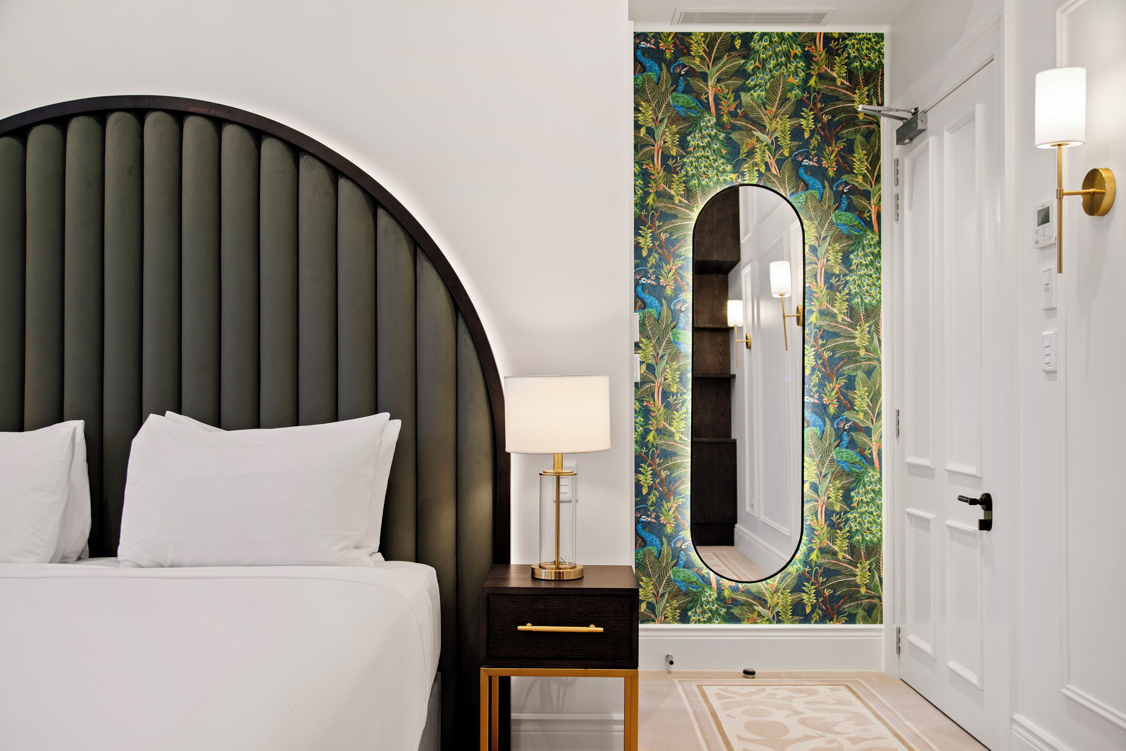 A bedroom with a large bed, a bedside table with a lamp, a mirror, and a door with a white wall and a decorative mirror.