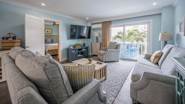 A living room with a sofa, armchairs, a coffee table, and a television. There is a balcony with a view of palm trees.