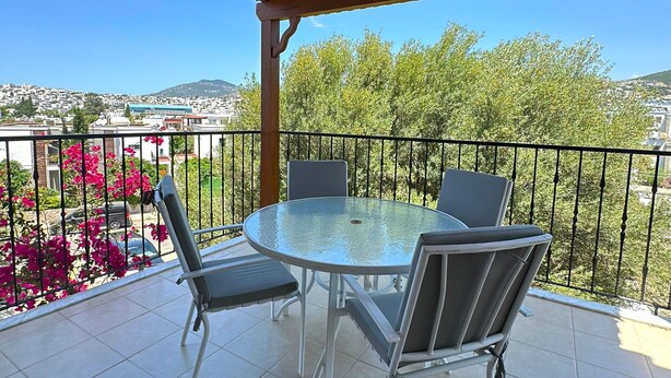 Une terrasse extérieure couverte, dotée d’une table ronde et de quatre chaises, qui offre une vue sur un quartier résidentiel, avec des arbres et une colline au loin.