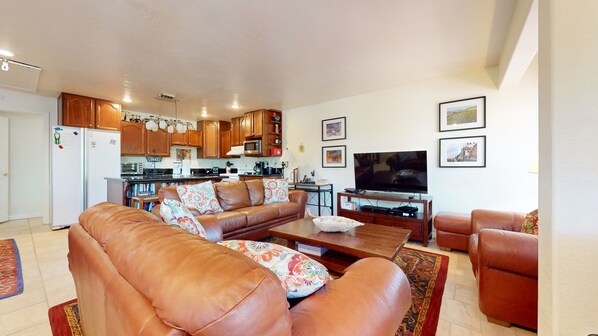 A living room with a brown leather sofa, a coffee table, and a TV. There is a kitchen in the background with wooden cabinets and a microwave. - Lakeview Villas #26 3 Bedroom Condo by RedAwning (Mammoth Lakes)