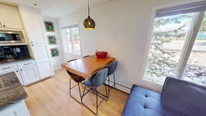A modern kitchen with white cabinets, a built-in microwave, and a coffee maker. There is a dining area with a wooden table, blue chairs, and a red basket. A large window offers a view of a rocky outdoor area. - Updated End Unit In Town! Walk to Coffee, Amp, Marina, Parks, Rec Path, Eats (Dillon)