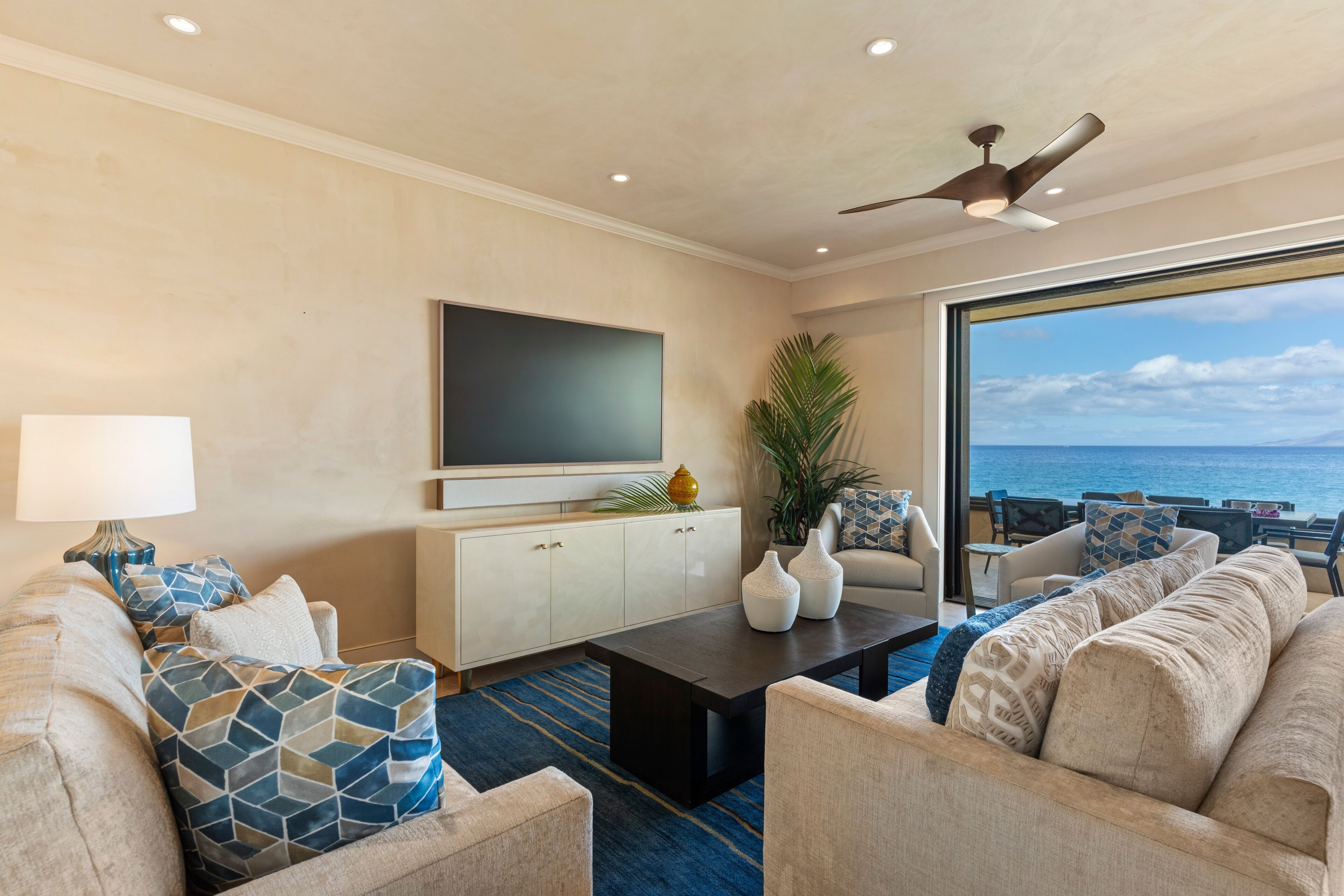 A modern living room with a flat-screen TV, a ceiling fan, and a large window overlooking the ocean.