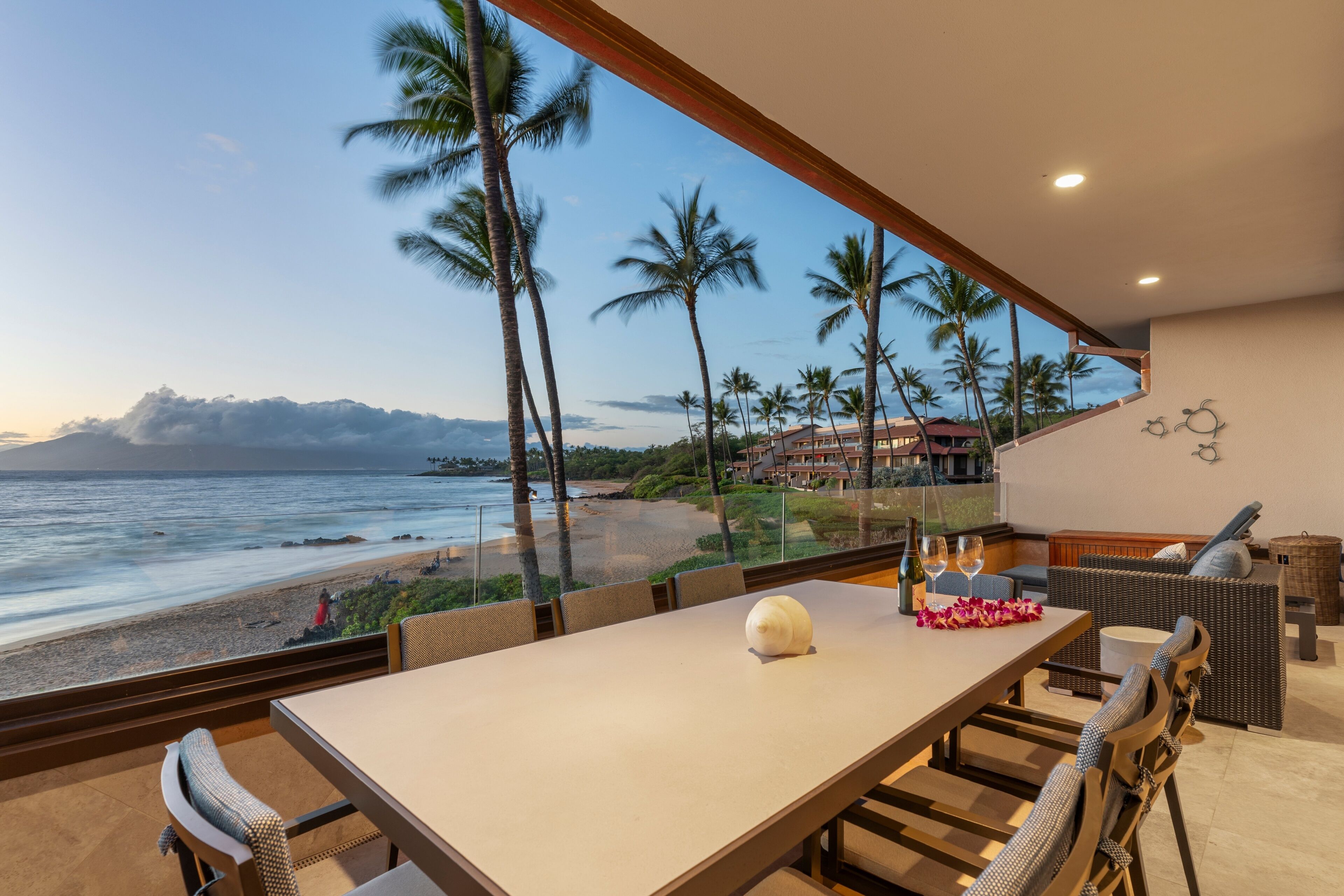 A dining area with a view of the beach and ocean, featuring a table with chairs, a decorative shell, and a bottle of wine.