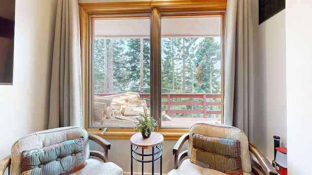 A room with two armchairs, a small round table with a plant, and a large window with a view of a deck and trees.