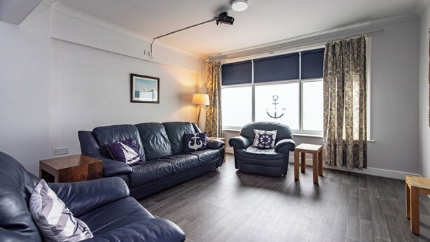 A modern living room with a dark leather sofa, a smaller armchair, and a wooden side table. A large window with curtains allows natural light to enter.