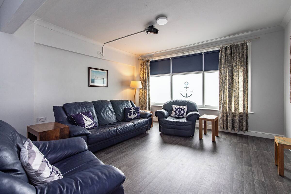 A modern living room with a dark leather sofa, a smaller armchair, and a wooden side table. A large window with curtains allows natural light to enter.