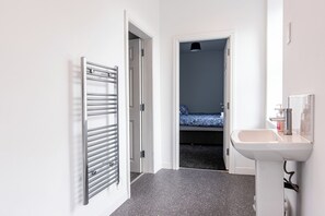 A hallway with a radiator, a bed with a blue bedspread, and a white sink. - Maritime House I (Hull)