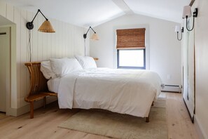 Un dormitorio con cama, un banco, dos lámparas de pared, una ventana con persianas y un radiador.
