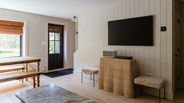 Una sala de estar con una mesa de madera, dos sillas, un televisor fijado a la pared y una puerta que da al exterior.