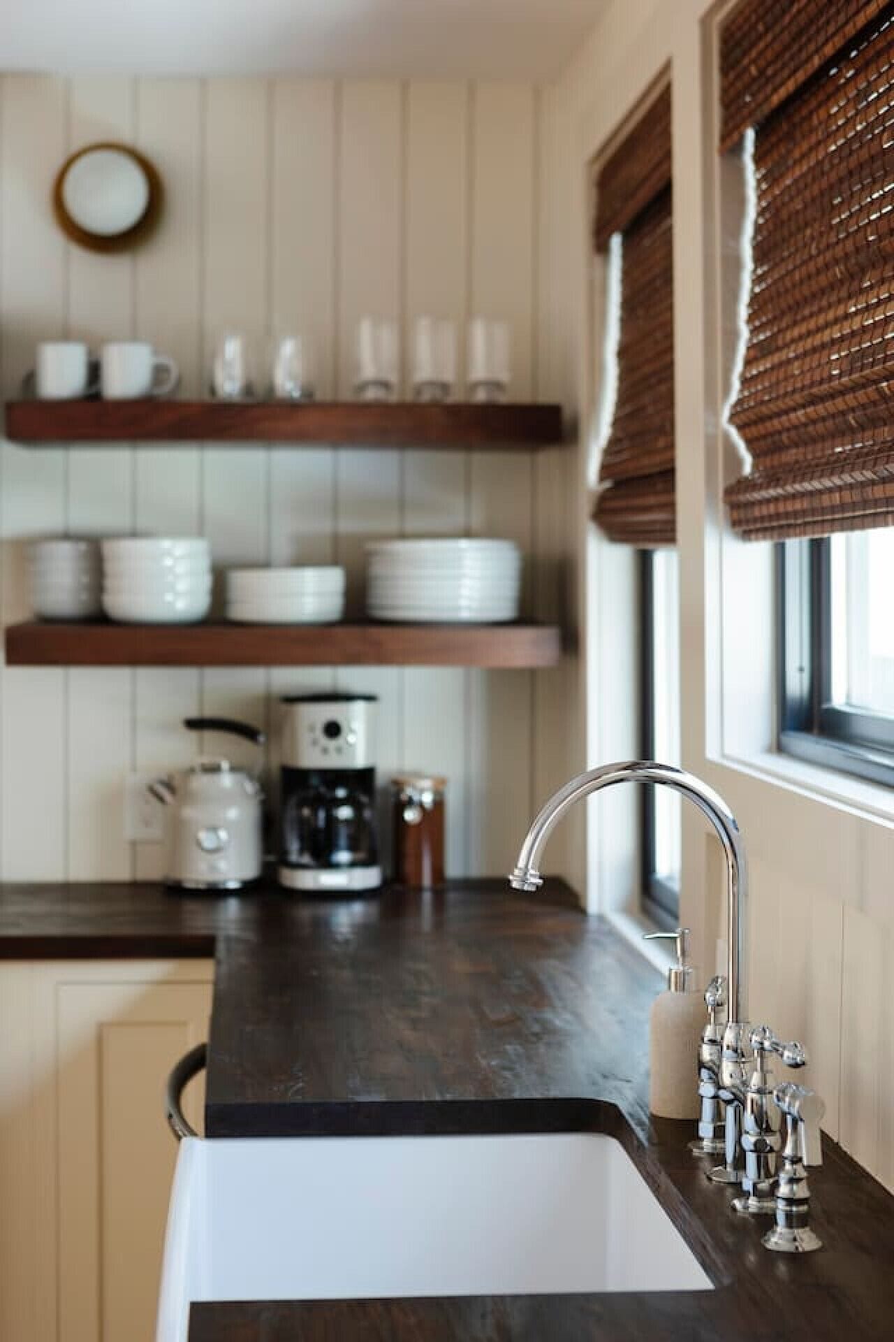 A kitchen with a dark countertop, a sink with chrome fixtures, and wooden shelves above holding white dishes and a coffee maker.