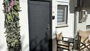 Un bâtiment blanc avec une porte noire, une petite fenêtre et un carillon. Il y a un paillasson noir devant la porte et une chaise à l’extérieur.