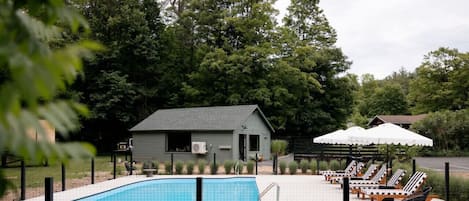 Una piscina con sillas de descanso y sombrillas, rodeada de árboles y un pequeño edificio.