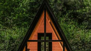 Una cabaña de madera triangular con un frente de vidrio, una banca y una mesa en su interior.