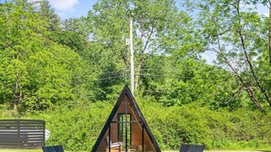 A cozy outdoor seating area with two Adirondack chairs and a fire pit, surrounded by lush greenery and a small wooden cabin.