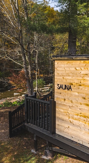 Une structure de sauna en bois, portant l’inscription « SAUNA », se trouve dans une zone boisée, et une passerelle en bois mène à cette structure.
