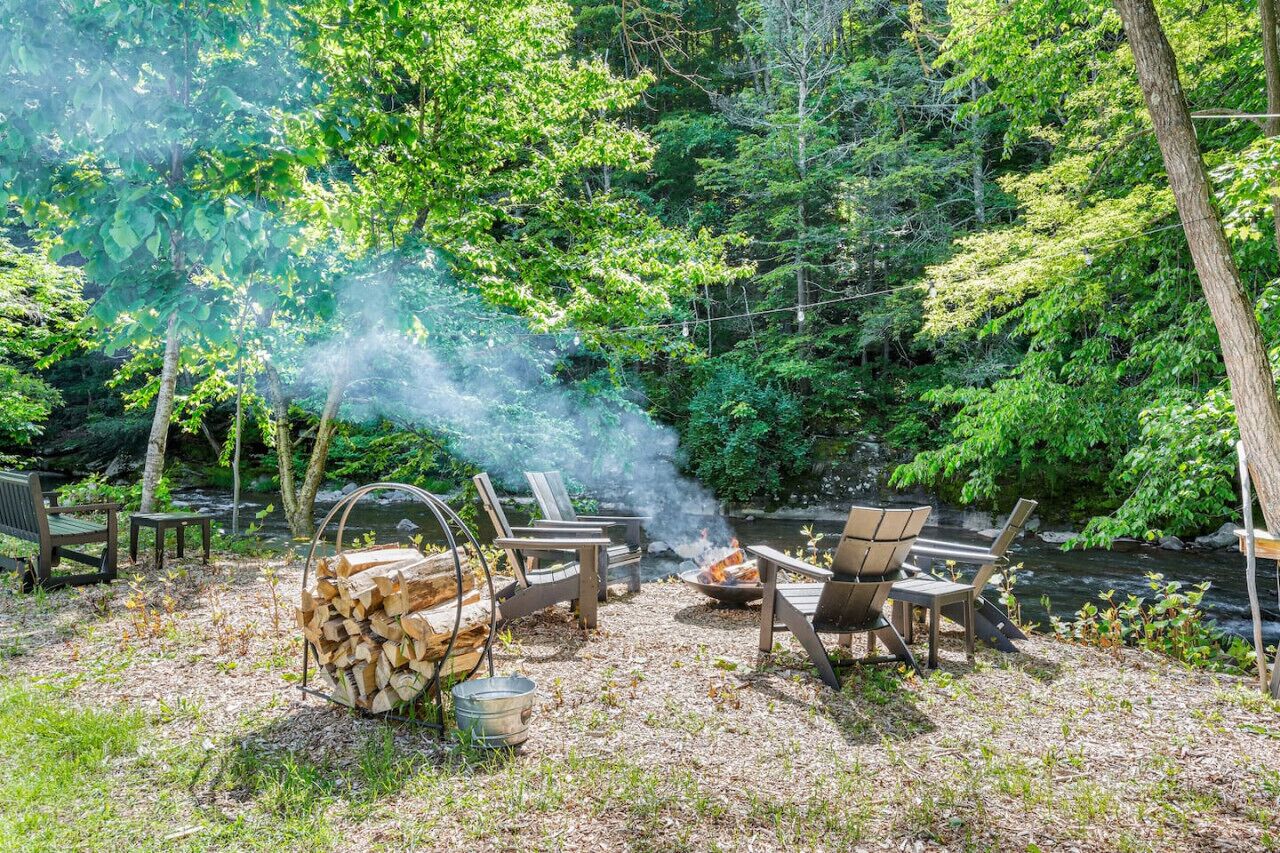 A forested campsite with a fire pit, chairs, and a stack of firewood.