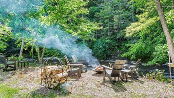 A forested campsite with a fire pit, chairs, and a stack of firewood.