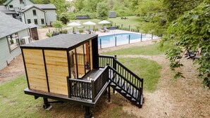 Una casita de juegos de madera con terraza y escaleras, una piscina y un jardín césped con árboles.