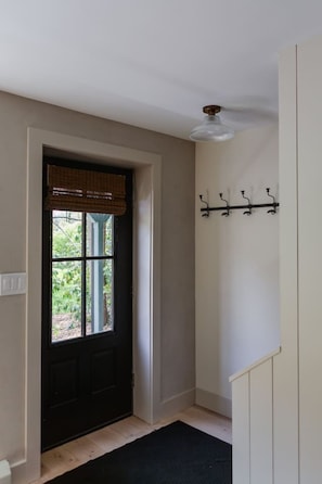 A hallway with a black door, a window with a view of greenery, a ceiling light fixture, and a coat rack with hooks.