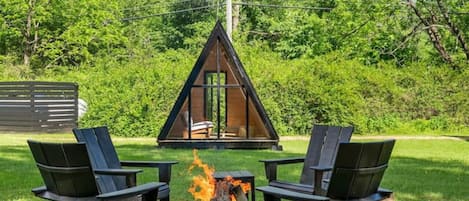 Ein gemütlicher Außenbereich mit zwei Adirondack-Stühlen und einer Feuerstelle, umgeben von üppigem Grün und einer kleinen Holzhütte.