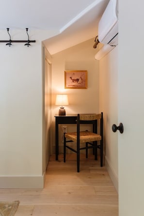 A narrow hallway with a wooden floor, a small table with a lamp, a chair, and a framed picture on the wall.