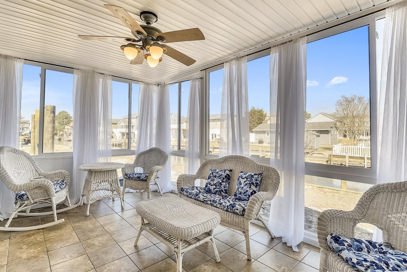 A sunroom with wicker furniture, a ceiling fan, and large windows offering a view of the neighborhood.