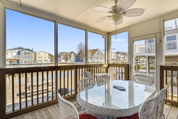 A sunlit room with a glass table, wicker chairs, a ceiling fan, and a view of neighboring houses.
