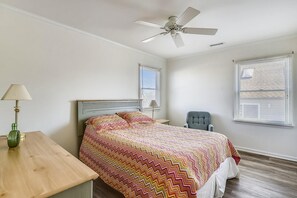 Quarto com cama, escrivaninha, cadeira, ventilador de teto e duas janelas com persianas.