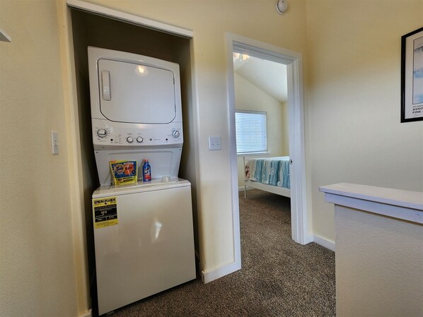 A laundry room with a front-loading washer and dryer, a small shelf with detergent bottles, and a doorway leading to a bedroom with a bed and a window. - Beachscape - Beautiful views (Ocean Shores)