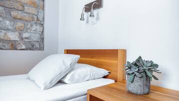 A neatly made bed with white linens and pillows, a wooden headboard, a small wooden nightstand with a potted plant, and a stone wall with a modern light fixture.