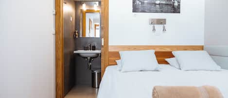 Habitación de hotel con cabecera de madera, cama con ropa blanca, toalla enrollada, lavamanos y espejo.