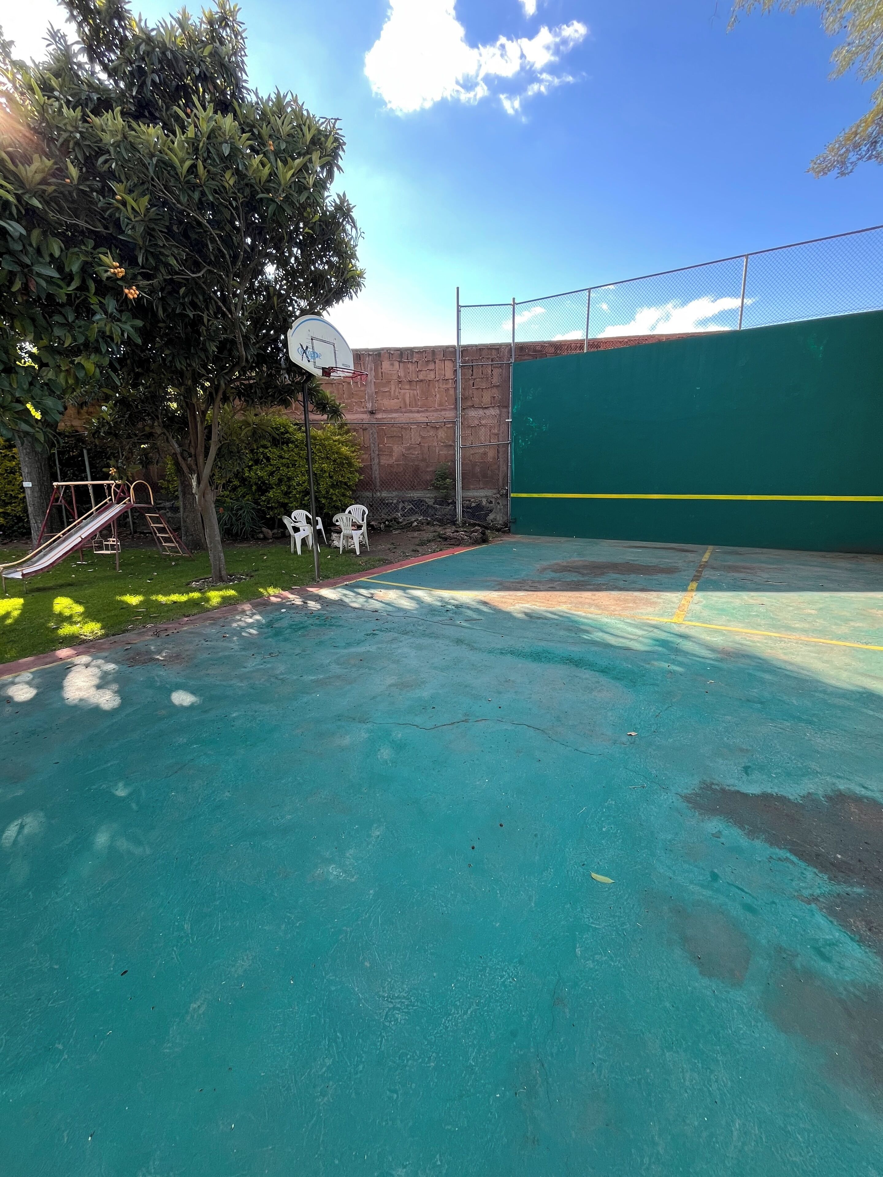 An outdoor basketball court with a green fence, a basketball hoop, and a slide.