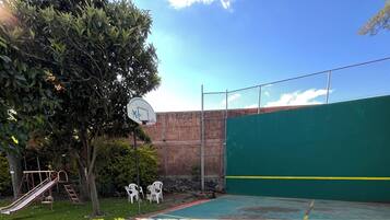 An outdoor basketball court with a green fence, a basketball hoop, and a slide.