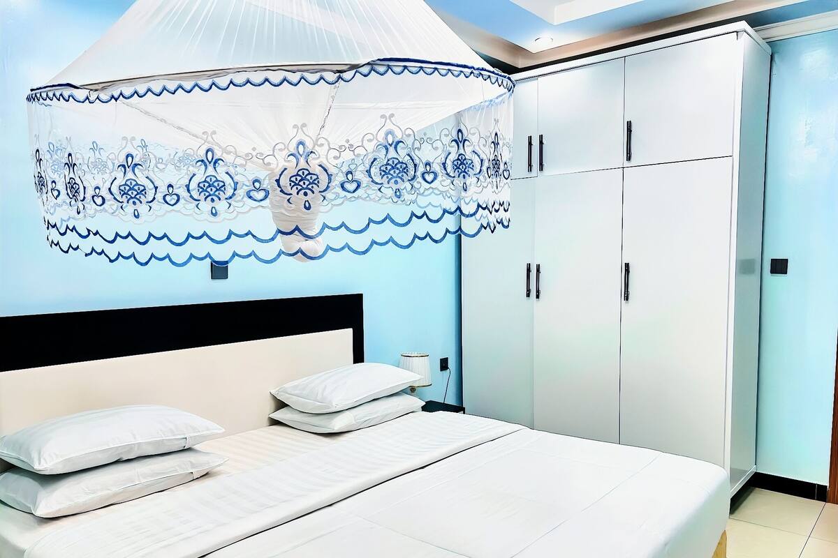 A bedroom with a bed, white bedding, a canopy with blue patterns, a wardrobe, and a nightstand.