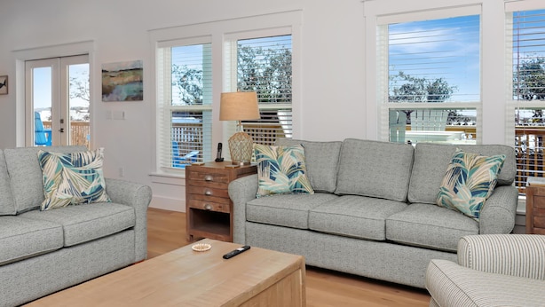 A living room with two grey sofas, a wooden coffee table, and a lamp. There are large windows with blinds, a painting on the wall, and a view of trees and a building outside. - Sacajawea by the Sea (Port St. Joe)