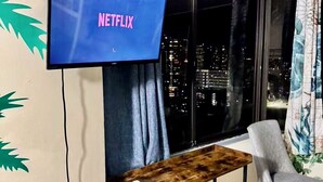 A room with a TV displaying 'NETFLIX', a wooden table with a shelf, a grey chair, and a window with a city view at night.