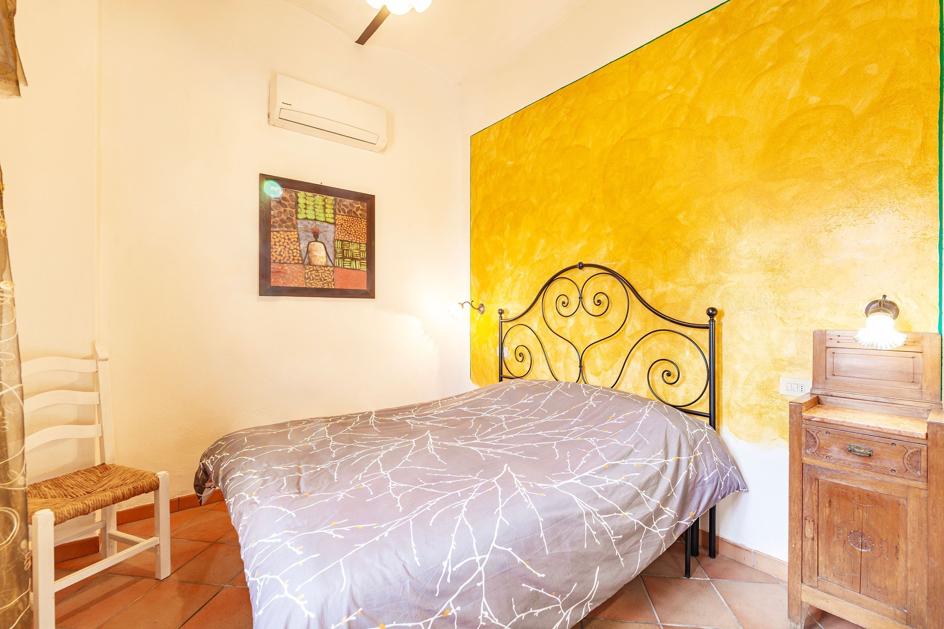 A bedroom with a bed, a chair, a wooden dresser, a wall with a yellow accent, and a framed picture on the wall.