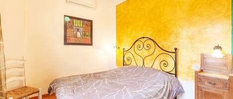 A bedroom with a bed, a chair, a wooden dresser, a wall with a yellow accent, and a framed picture on the wall.