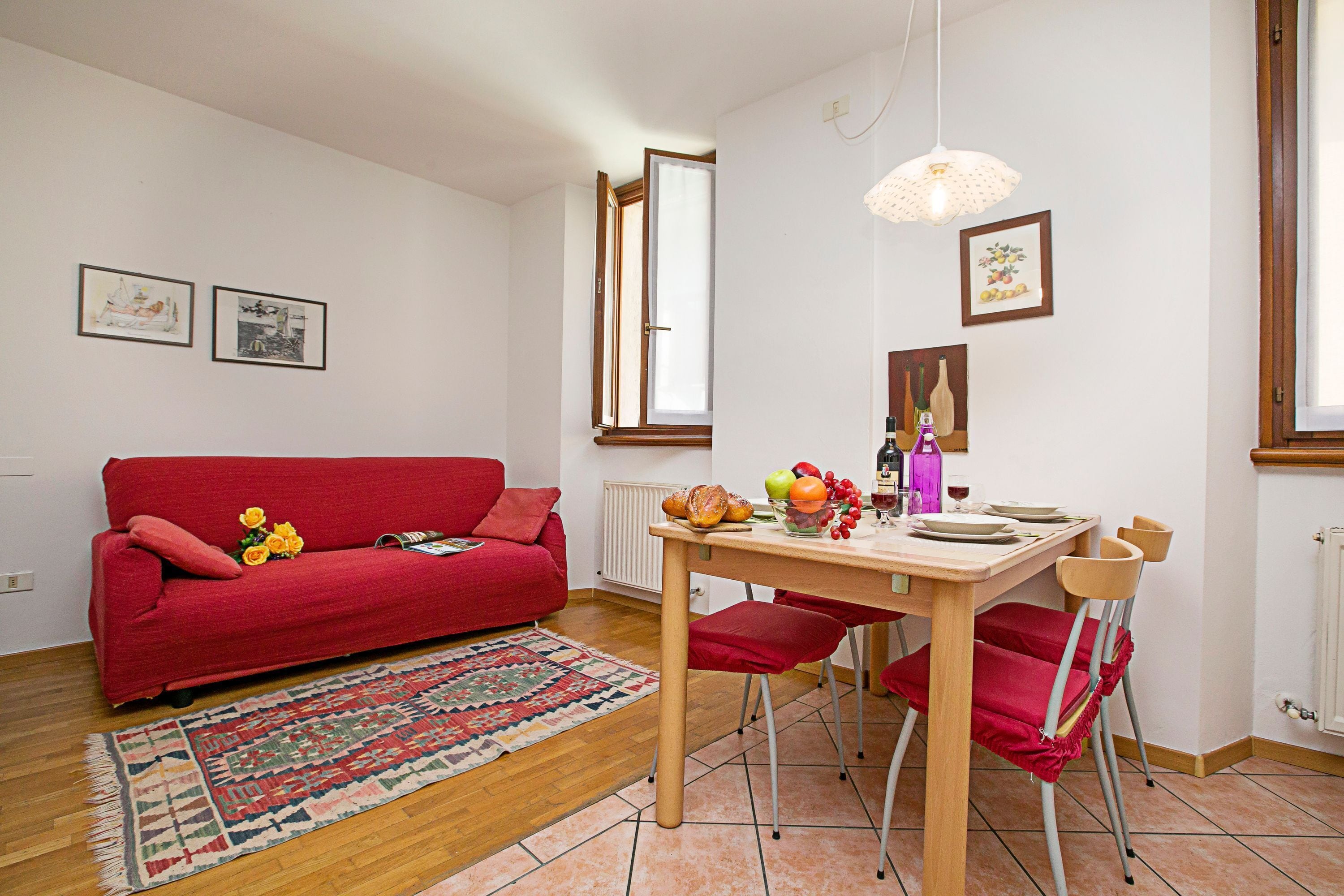 Una sala de estar con un sofá rojo, una mesa de comedor de madera con sillas, una alfombra y una ventana con cortinas.