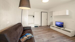 Ein modernes Wohnzimmer mit einer Couch, einem Fernseher auf einem weißen Schrank, einer Lampe und einer Tür im Hintergrund.