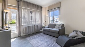 Een moderne woonkamer met een grijze bank, een kamerkleed met patroon en een groot raam met gordijnen.