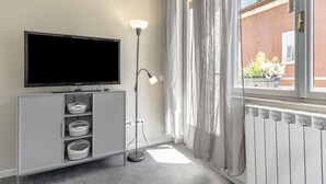 Een moderne woonkamer met een flatscreen televisie op een grijze kast, een vloerlamp en uitzicht op een gebouw van rode bakstenen door een raam met dunne gordijnen.